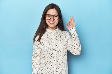 Young Caucasian woman on blue backdrop winks an eye and holds an okay gesture with hand.