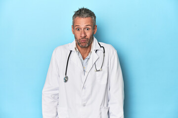 Caucasian middle-aged doctor on blue background shrugs shoulders and open eyes confused.