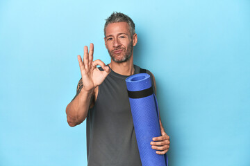 Athlete with a mat on a blue studio backdrop cheerful and confident showing ok gesture.