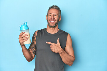 Athlete holding protein shake on blue backdrop smiling and pointing aside, showing something at...