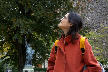 Caucasian woman in her 30s or 40s looking up daydreaming with orange coat in autumn with a copy of liberty statue in the background