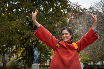 Caucasian woman in her 30s or 40s with black hair and orange coat  in Paris in autumn making victory gesture very happy, looking at camera smiling