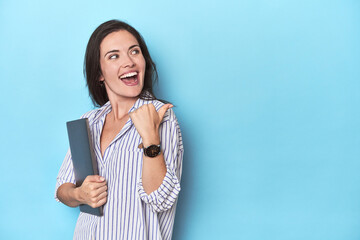 Young businesswoman holding laptop on blue points with thumb finger away, laughing and carefree.