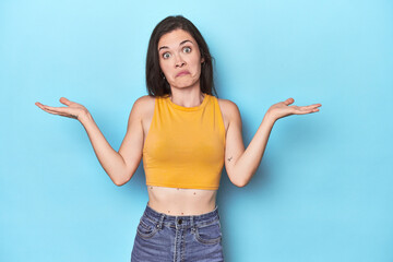 Young caucasian woman on blue backdrop doubting and shrugging shoulders in questioning gesture.