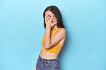Young caucasian woman on blue backdrop blink at the camera through fingers, embarrassed covering...