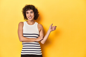 Caucasian curly-haired woman in white tank-top smiling cheerfully pointing with forefinger away.