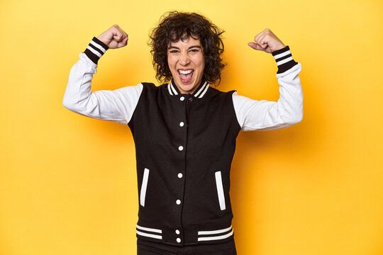 Curly-haired Caucasian woman in baseball jacket showing strength gesture with arms, symbol of feminine power