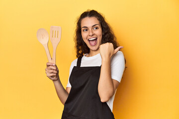 Woman with apron, wooden cooking utensils, yellow, points with thumb finger away, laughing and carefree.