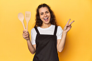 Woman with apron, wooden cooking utensils, yellow, joyful and carefree showing a peace symbol with fingers.