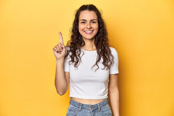 Young Caucasian woman, yellow studio background, showing number one with finger.