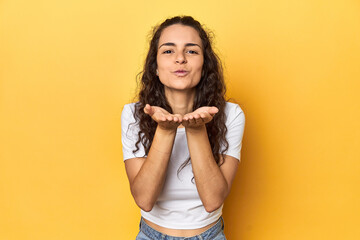 Young Caucasian woman, yellow studio background, folding lips and holding palms to send air kiss.