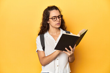 Young Caucasian woman engrossed in reading a book in a studio with yellow backdrop.