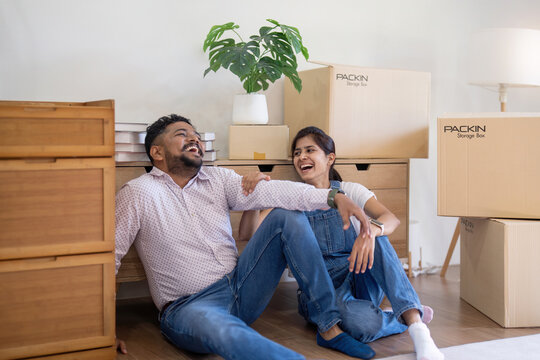 Happy young indian couple taking a break on moving day into new home sitting on floor surrounded by box