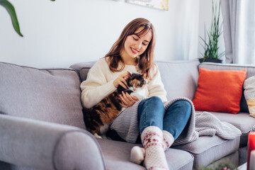 Obraz premium Woman in plaid with tea cup watching movie, TV, petting cat on sofa at home with christmas decoration atmosphere. Lady wear jumper and warm socks. Cozy and comfortable winter concept. Selective focus.
