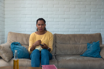Young scared woman sitting alone at home on the sofa watching scary horror movie on the television....