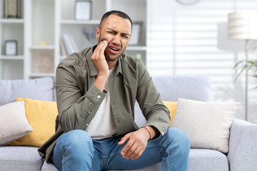 Young hispanic man suffering from toothache sitting on sofa at home and holding hand to cheek