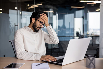 Upset young Indian male businessman sitting at desk in office, looking upset at laptop screen and...