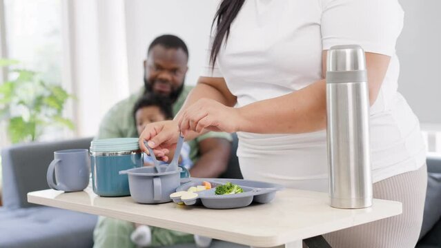 Asian mother prepares food for feeding her 9 months old her cute little baby and African American holds the baby on the sofa. Photo series of family, kids and happy people concept. Parents feed kids.