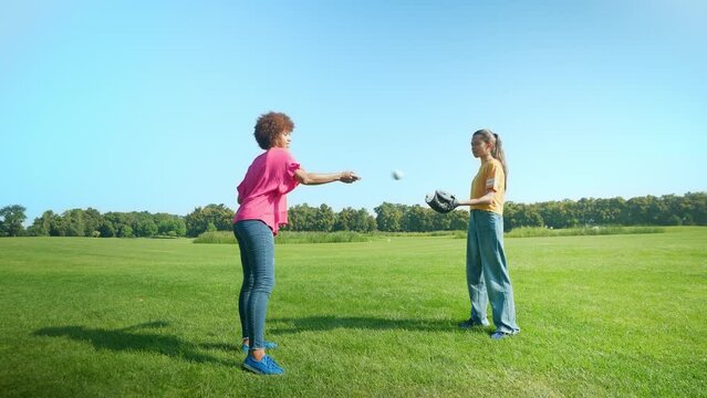 Positive attractive black mother and happy cute multiracial teenage daughter in gloves having fun, playing and throwing baseball ball on green field while family enjoying leisure in summer nature.