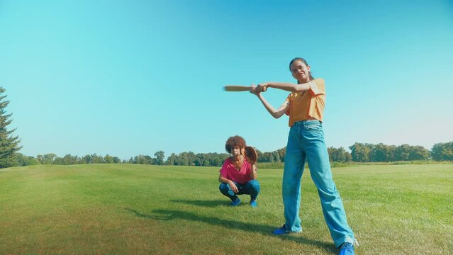 Active adorable multiethnic teenage girl baseball player with positive black mother catcher with glove playing baseball game, batting pitch on green field while family spending leisure in nature