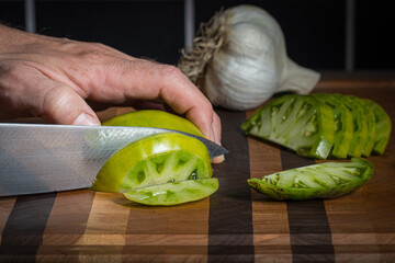 A man using a knife to slice a Cherokee Green Tomato while making salsa with a head of garlic in the background.
- 650687894