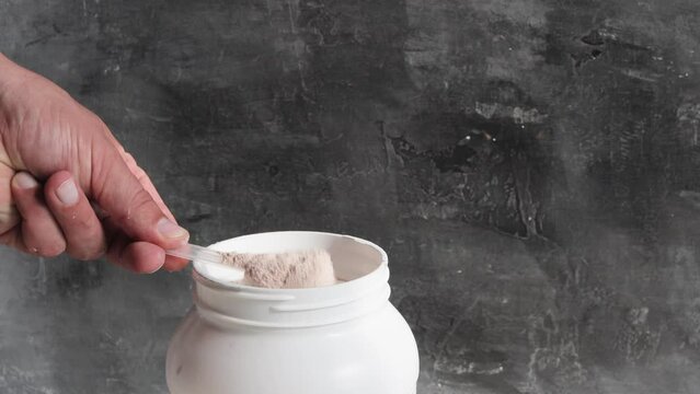 Male hand with measuring scoop takes out chocolate whey protein powder from plastic jar on dark background, close-up view