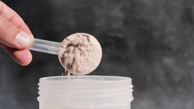 Male hand puts into a shaker portion of chocolate whey protein powder from measuring scoop on dark background, slow motion, close-up view