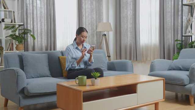 Upset Asian Woman Using Smartphone While Sitting On Sofa In The Living Room 
