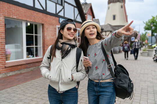excited asian chinese girl backpacker showing friend a famous landmark in distance with pointing hand gesture during their stay in solvang California's Danish village in usa