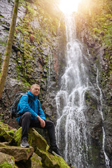 Obraz premium Tourist attraction of Germany - falls of Burgbach Waterfall near Schapbach, Black Forest, Baden-Wurttemberg, Germany. Man hiker in blue jacket standing on stone and looks at flow of falling water