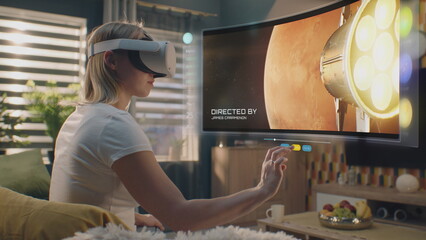 Woman in VR headset chooses movie or TV show to watch at home. 3D futuristic hologram shows...