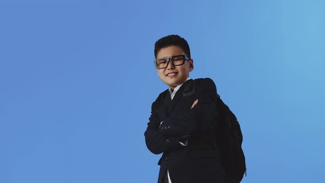 Young asian boy in glasses and school uniform with a backpack crosses hands on a chest and confidently smiles on a blue background. Concept of modern interesting education for children