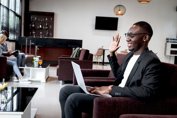 African businessman working on laptop computer while sitting on sofa in airport business lounge or hotel lobby, happy smiling male has online conference meeting and waving hand for greeting.