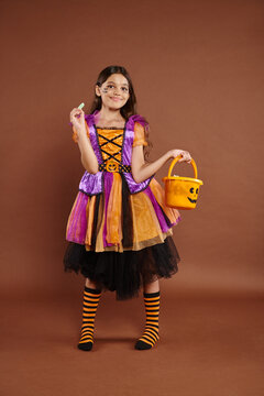 lovely girl in Halloween costume holding bucket and looking at wrapped candy on brown backdrop