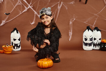 cheerful preteen girl squatting near pumpkin with lanterns and spiderweb on backdrop, Halloween