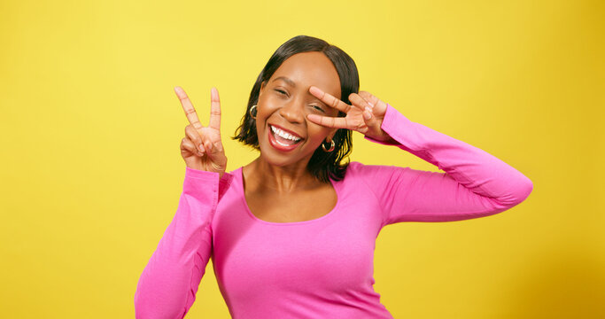Happy young woman holds peace sign to eye, silly pose, yellow studio portrait