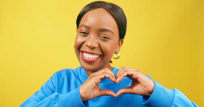 Young Black woman forms heart with hands, smiling yellow studio background