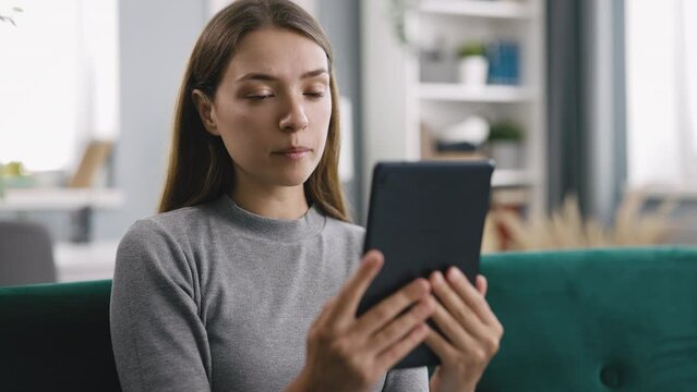 Young woman having video chat from home, sitting on couch in brightly lit living room, spends free time conversating with friend or family