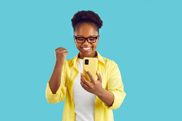 Joyful excited lucky woman with mobile phone in hands is happy about good news or win. Happy young african american woman with smartphone joyfully clenches fist against light blue background.
