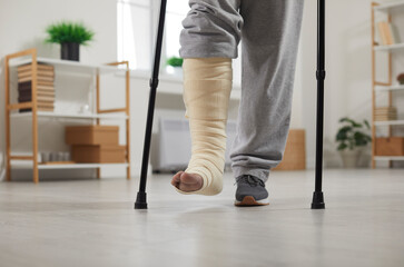 Man with fractured leg in cast standing with crutches at home, selective focus. Cropped view of plastered leg and crutches. Bone fracture, injury, trauma, recovery, rehabilitation concept - obrazy, fototapety, plakaty