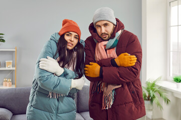 People get frozen inside a very cold house with heating problems. Family couple wearing warm winter clothes at home. Husband and wife in coats, hats and gloves looking at the camera and shivering