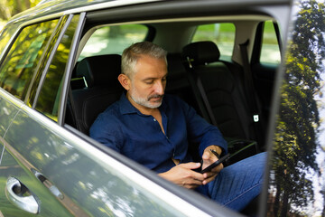 Handsome man in suit is using a smart phone and smiling while sitting on back seat in the car