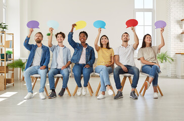 Group of happy young diverse multiracial people holding up colorful paper mockup speech bubbles while sitting in row on chairs in new modern office or rented apartment. Opinion survey concept