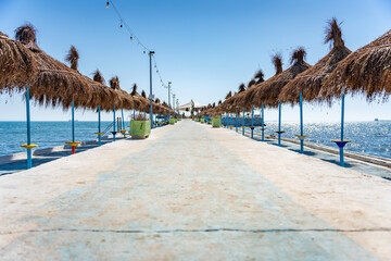 Walkway along a path of tiki umbrellas and ocean water on both sides - obrazy, fototapety, plakaty