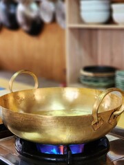 Brass wok sit on a gas stove with flames and a kitchen backdrop with cooking utensils and a plate rack.
