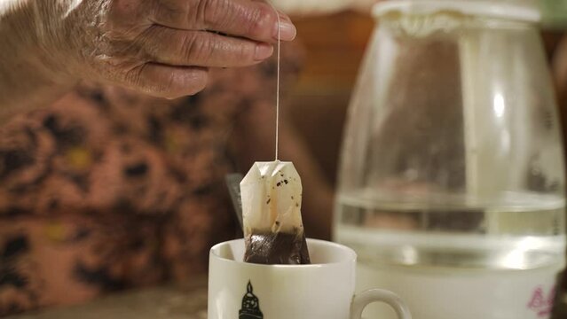 An elderly woman brews tea bag in white mug close-up. Grandma is having breakfast