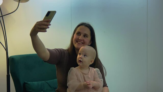 Young Caucasian mother making selfie with her pretty baby daughter. Using gadgets for social media. Woman taking pictures of her toddler and herself