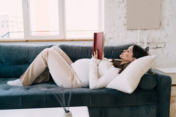 Young pregnant woman lying on sofa and reading book in living room