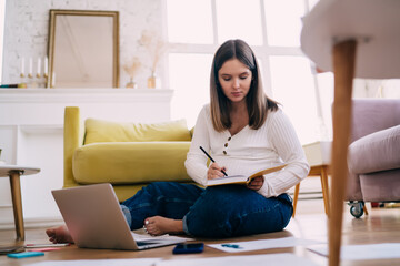 Pregnant woman writing in notebook on floor near laptop