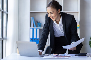 Young asian businesswoman sitting at desk with data and graph papers by using laptop at the office workplace, Accounting analyze, plan, and strategize about business improvement or finance.